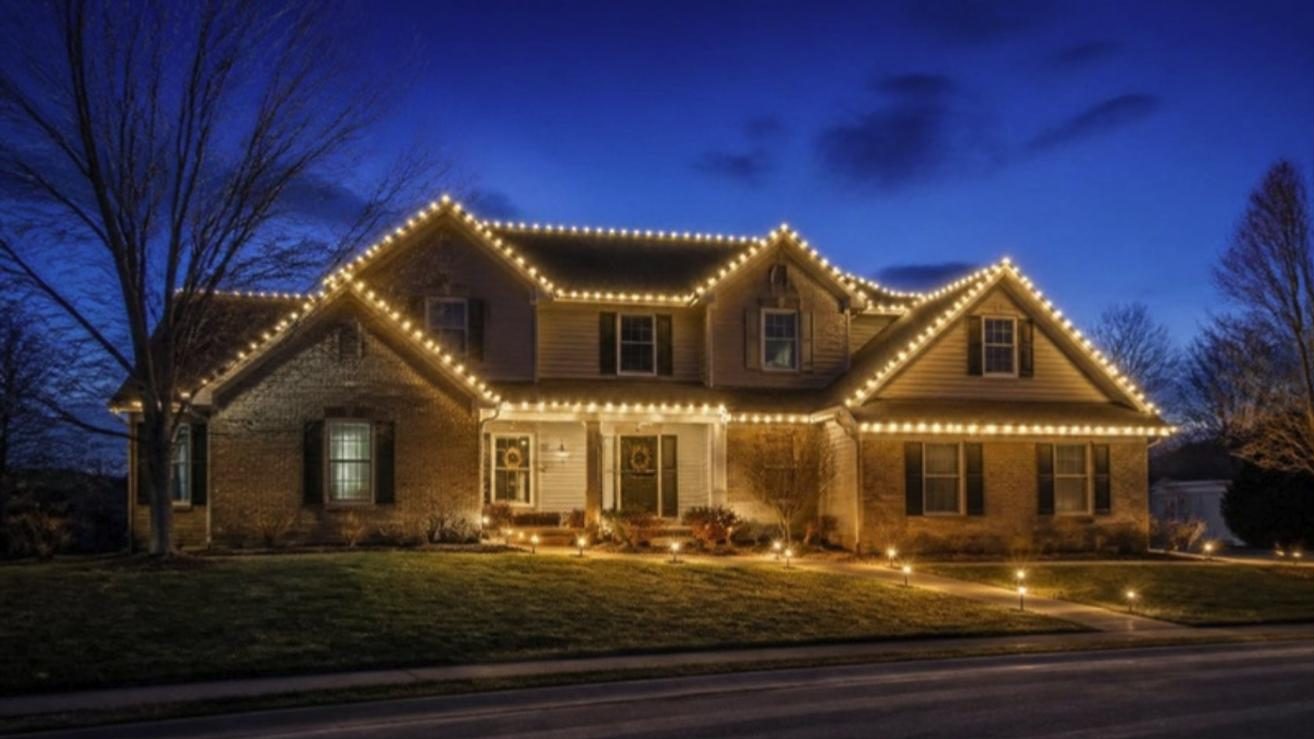 Two-story brick home at dusk with every roofline outlined in warm white Christmas lights